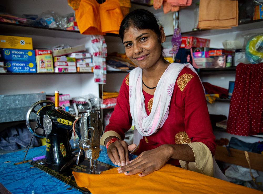 A woman smiles while sewing at her machine, surrounded by supplies.