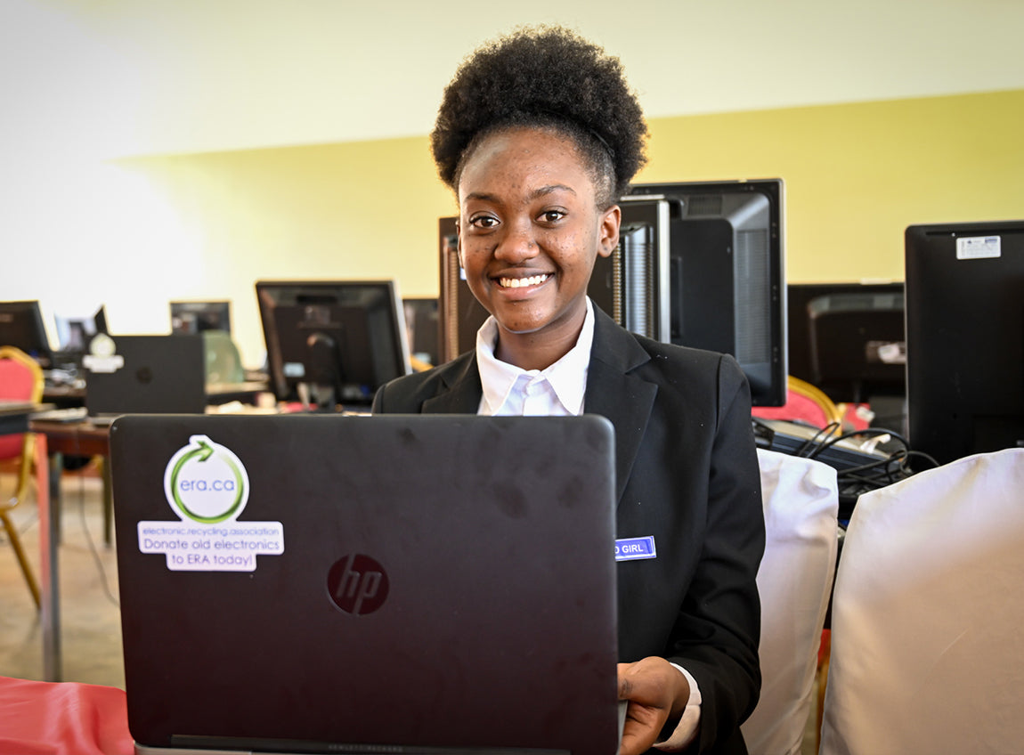 A girl smiles to the camera while sitting in a computer lab with a laptop in front of her.