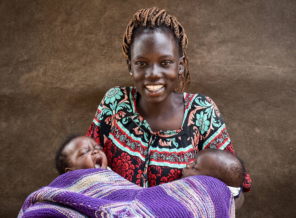 A woman smiles for the camera as she cradles two newborns wrapped in a purple blanket.