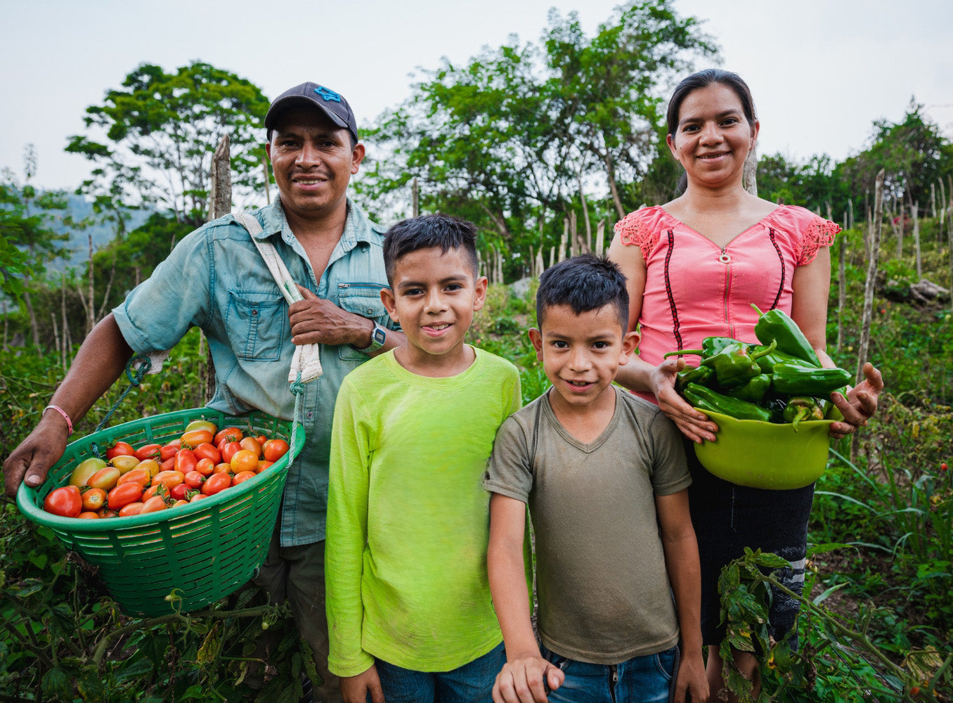 A family stands together in a garden, smiling. The father holds a basket of tomatoes, the mother holds a bowl of green bell peppers, and their two sons stand in front wearing bright t-shirts. The background is filled with greenery.