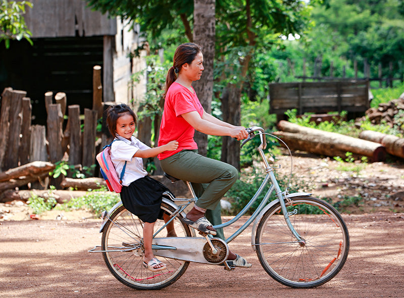 A woman rides a bicycle with a smiling child sitting behind her. The child is wearing a school uniform and backpack. The background shows a rural setting with trees and wooden structures.