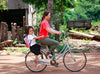 A woman rides a bicycle with a smiling child sitting behind her. The child is wearing a school uniform and backpack. The background shows a rural setting with trees and wooden structures.