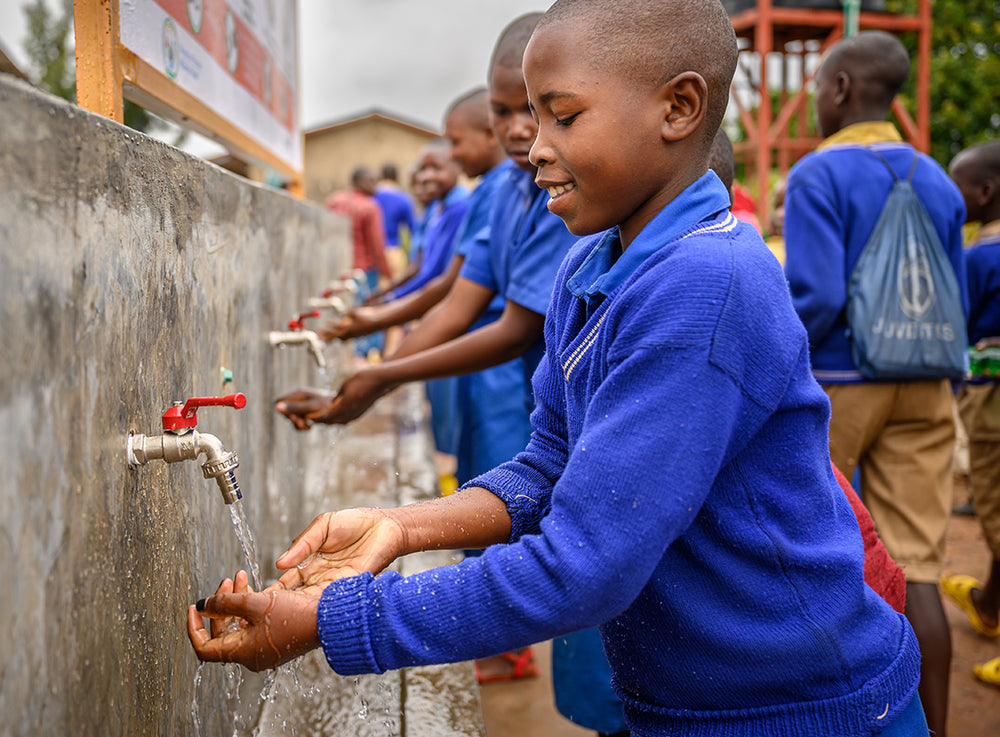 School children dressed in blue sweaters gather by a communal sink, washing their hands.