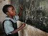A young girl in a classroom points to the alphabet on a chalkboard.