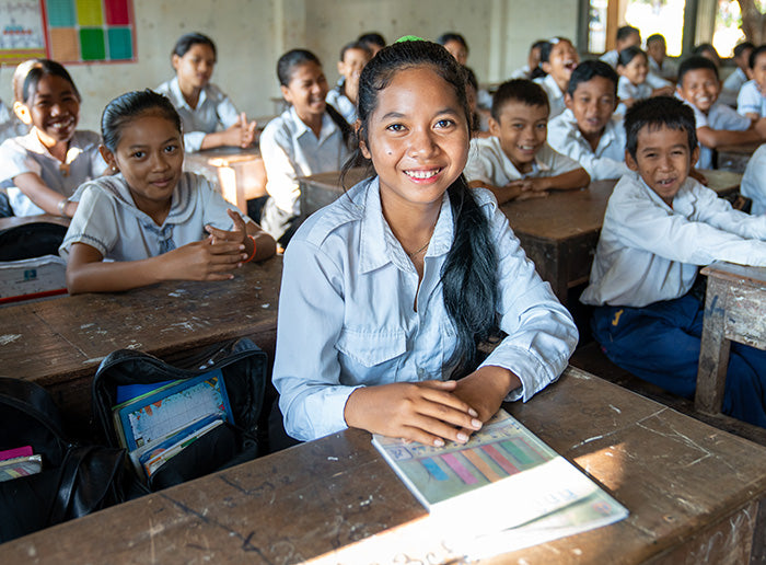 A classroom of smiling students 
in uniforms sit at wooden desks, with one girl prominently in the foreground holding a book.