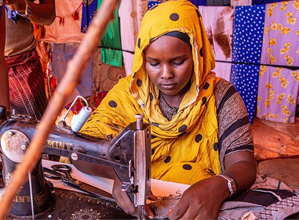 A woman wearing a yellow headscarf works at a sewing machine, surrounded by colorful fabrics.