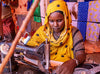 A woman wearing a yellow headscarf works at a sewing machine, surrounded by colorful fabrics.