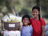 A sister places her hand over her brother's shoulders as they both smile while standing next to essential food items provided by World Vision.