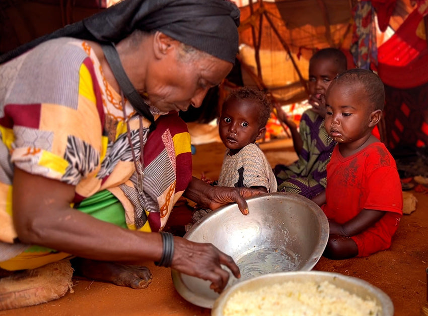 A grandmother serves food from a large bowl to her three grandchildren sitting on the ground, inside a tent-like structure.