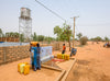 People filling containers with water from a community tap in a dry, rural area, with a water tower in the background.