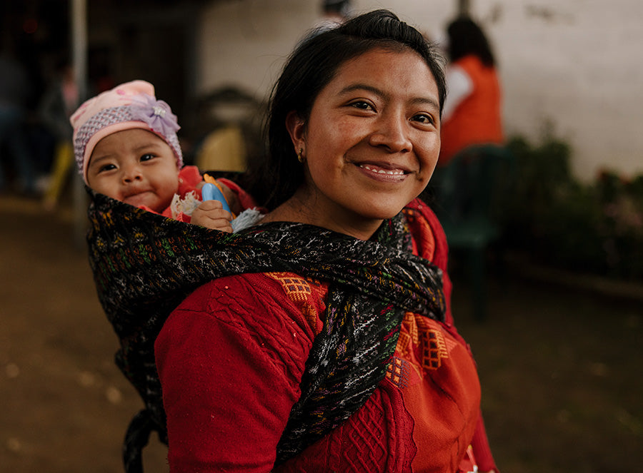 A woman wearing a red shirt with a baby wrapped around her back smiles as the  baby peeks over her shoulder.