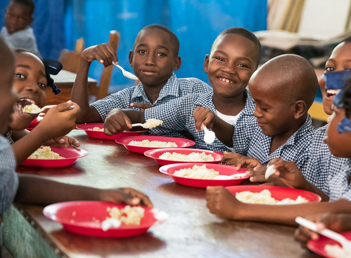 A group of school children eat lunch while smiling towards the camera.