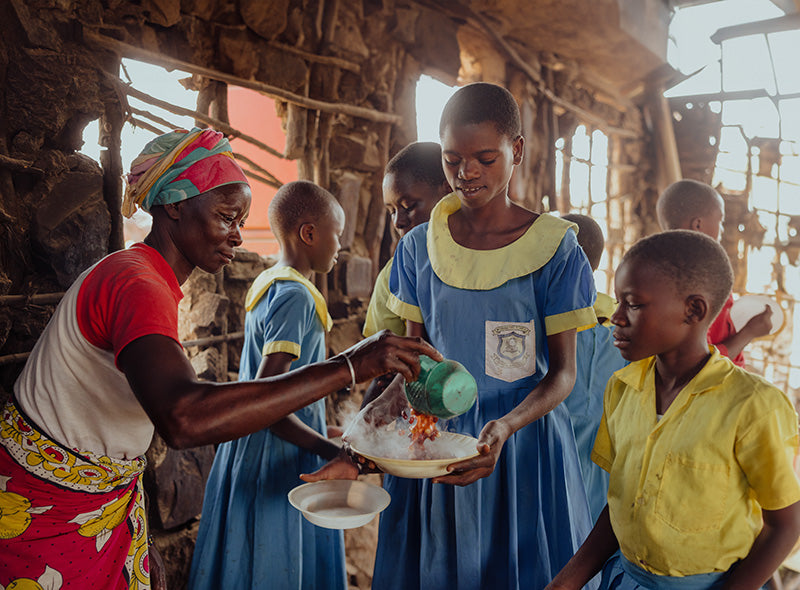 A woman in a red and white shirt with a colorful headscarf serves food to schoolchildren dressed in blue and yellow uniforms. 