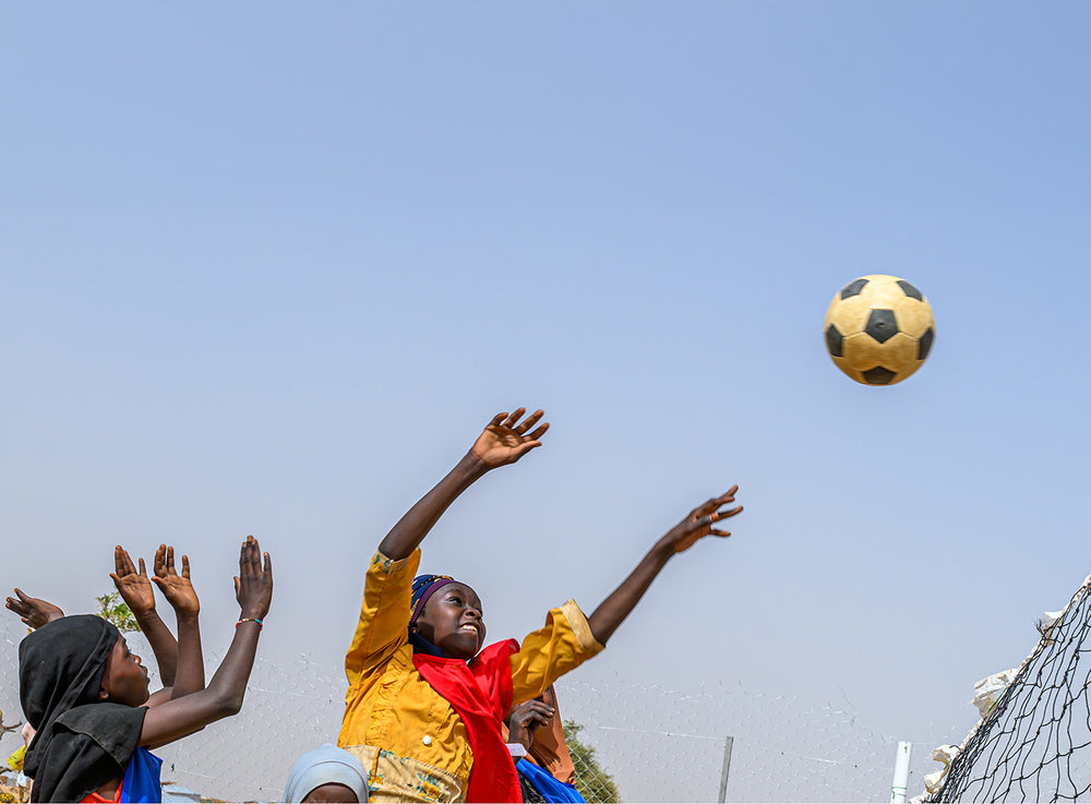 A group of children excitedly reach up toward a soccer ball in mid-air near a goal net, under a clear blue sky.