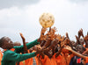 A large group of children reach their arms out towards a soccer ball in the air.
