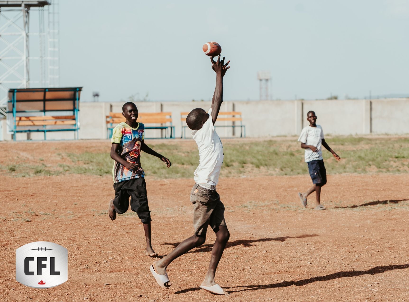 A group of children playing excitedly reach up toward a football. The Canadian Football League logo is overlaid on the left bottom corner of the image.