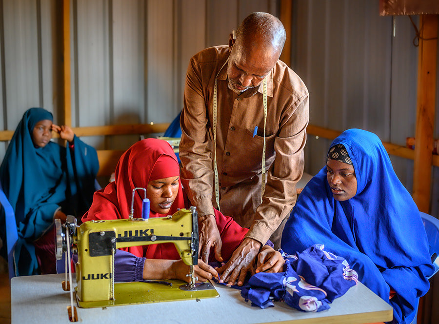 A man instructing a woman using a sewing machine while two other women observe, inside a workshop.