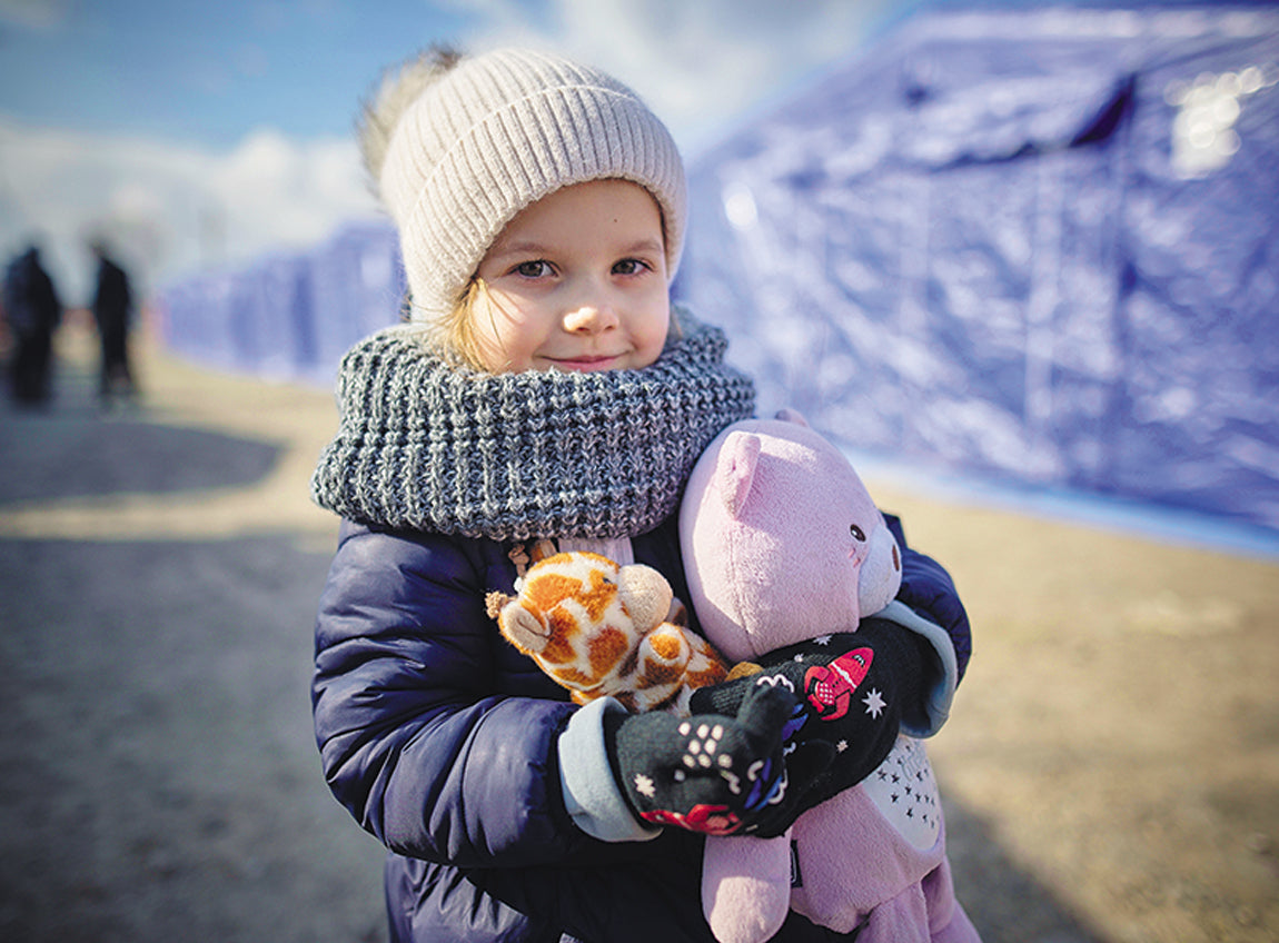 A young girl stands in front of refugee tents, dressed in warm clothing while clutching stuffed animals.