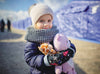 A young girl stands in front of refugee tents, dressed in warm clothing while clutching stuffed animals.