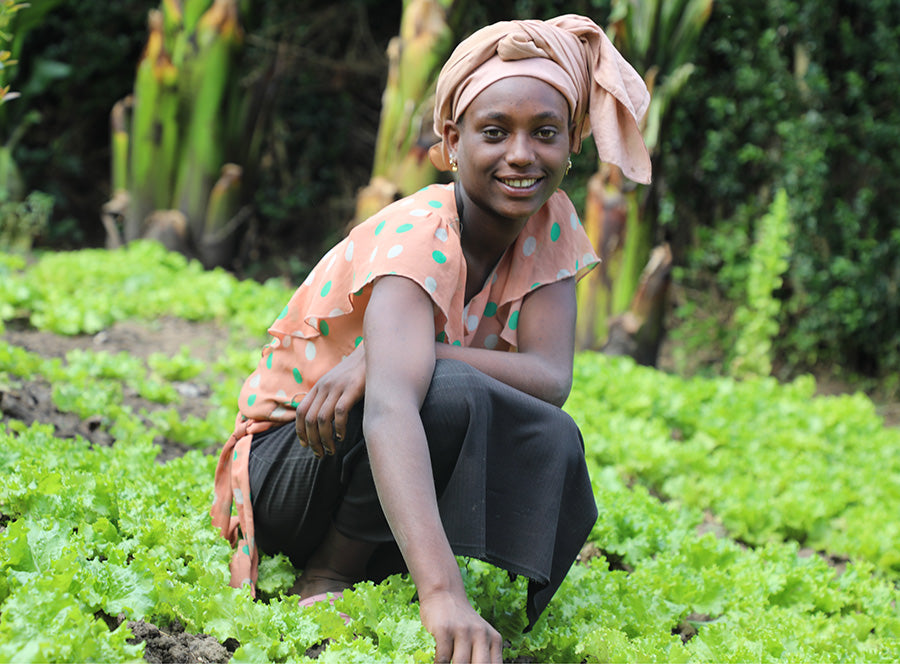 A woman smiles as she squats on an area surrounded by greenery.