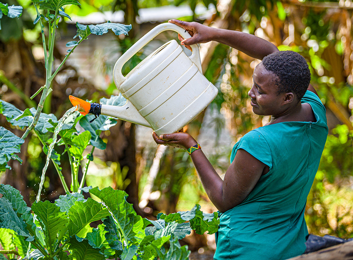 A woman holds a large watering can in the air, watering lush plants.
