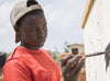 A teenage girl in an orange shirt and a grey cap worn backward is painting a mural.