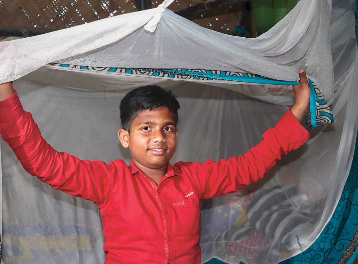 A boy lifts one side of a white-and-blue bed net hanging in his home.