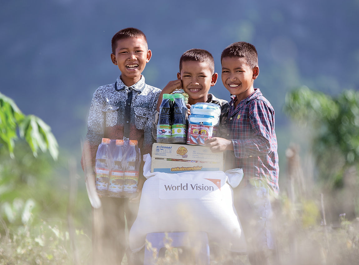 Three boys smile next to each other as they stand behind a pile of food items.