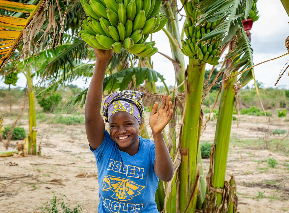 A young woman smiles and waves to the camera while grabbing a bushel of bananas in a tree.