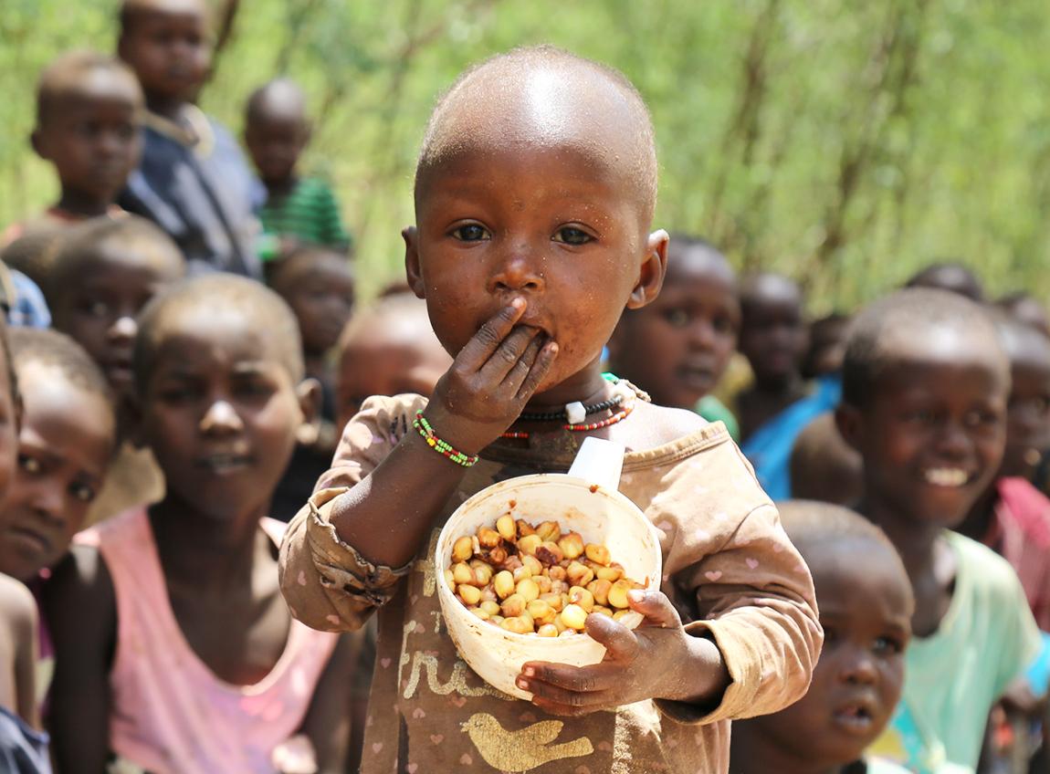 A very young child looks into the camera as he eats food from a container in his hands. Several more children sit in the background.
