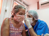 A healthcare worker wearing protective face, hand and head gear, examines the ear of a female patient, also wearing a mask.