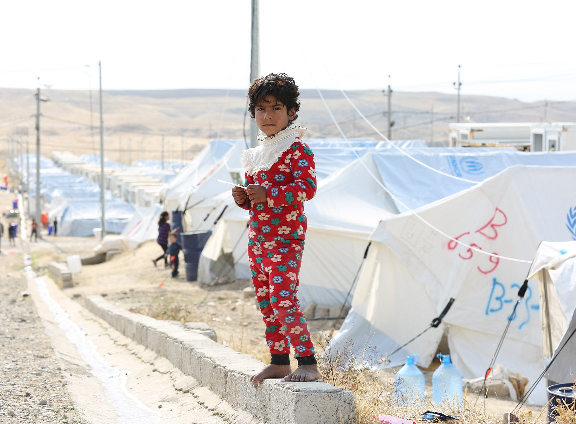 A yound girl dressed in floral pajamas, stands on a concrete wall overlooking tents in a refugee camp.