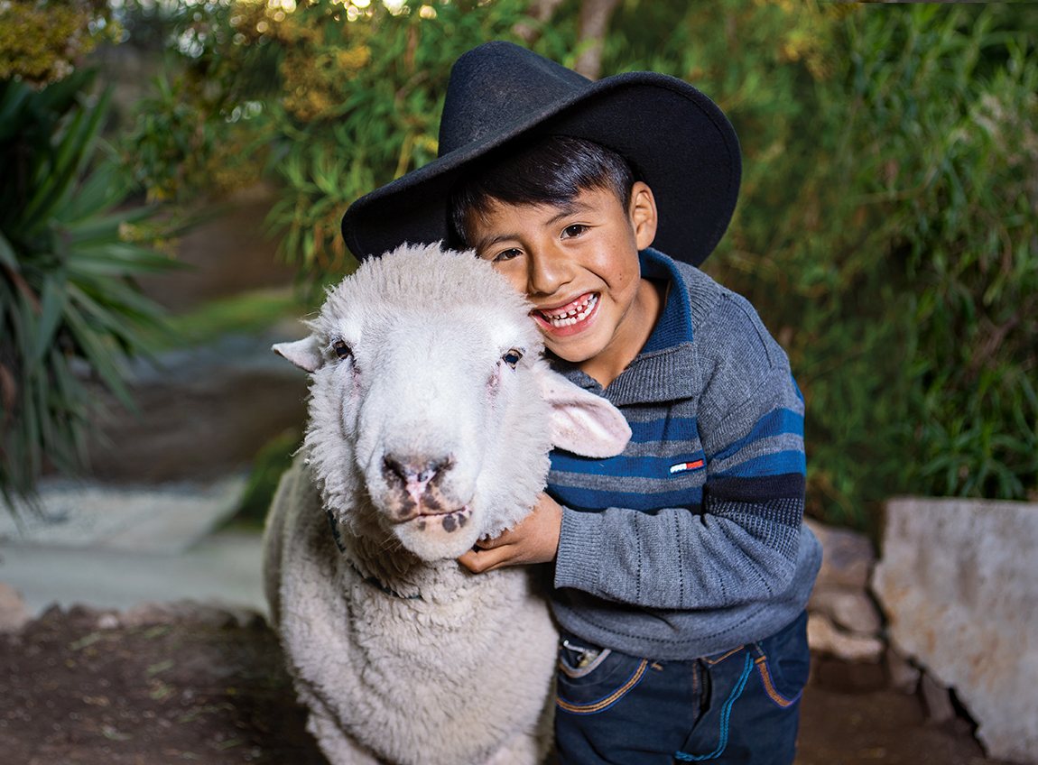 A smiling young boy wearing a black cowboy hat embraces a sheep.