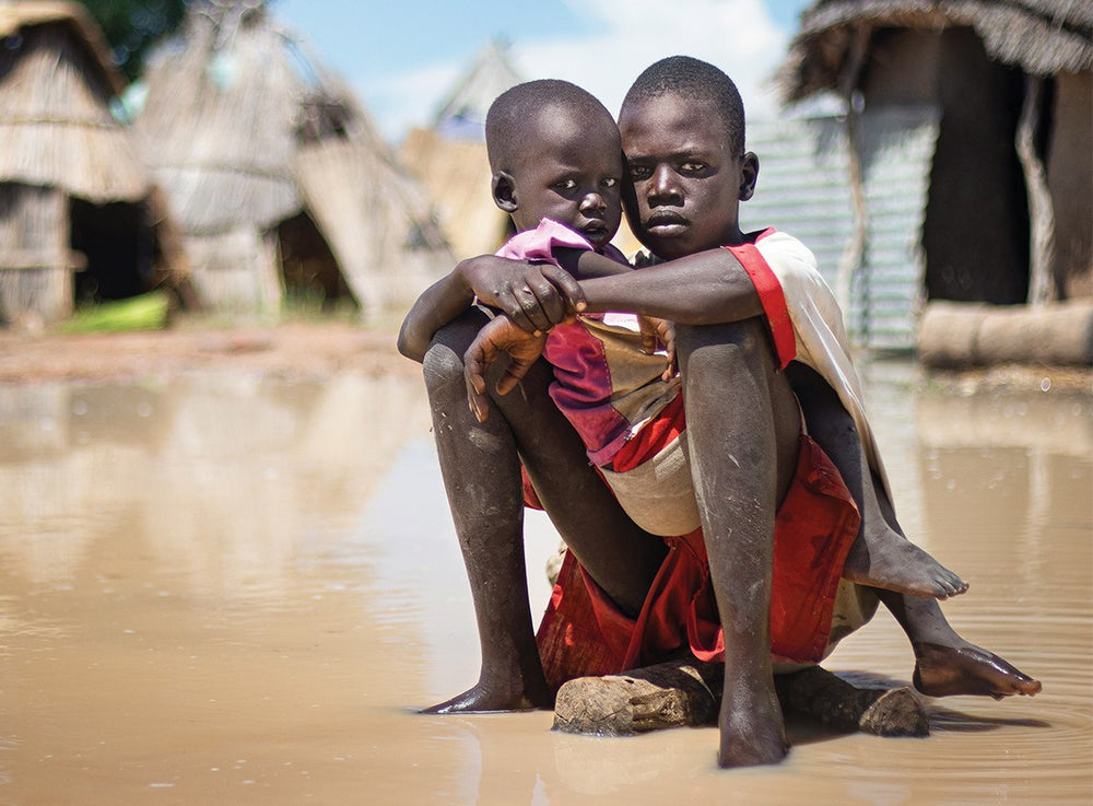 Two children with stern faces look into the camera as they sit on the ground near a group of huts.