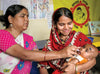 A young woman smiles as the baby she’s carrying receives oral vaccine from another woman.